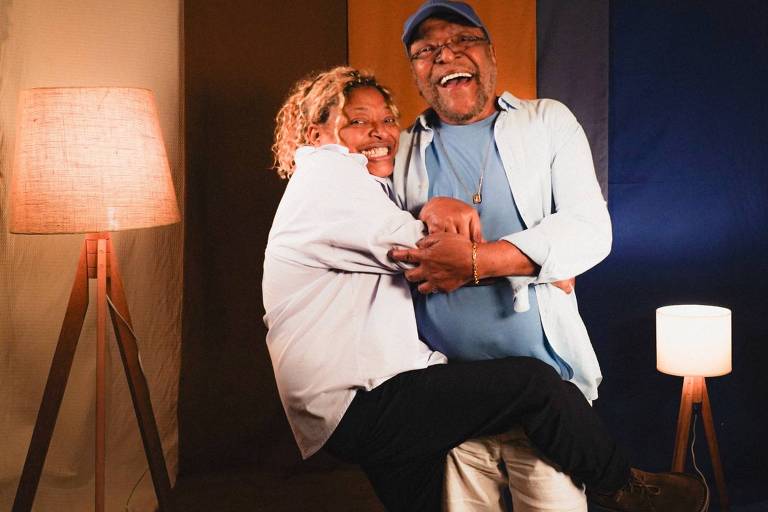 A imagem mostra uma mulher e um homem sorridentes em um ambiente interno. A mulher, com cabelo cacheado e loiro, está sendo segurada pelo homem, que usa um boné azul e óculos. Ambos estão rindo e se divertindo. Ao fundo, há painéis de tecido em cores variadas, e duas luminárias de pé, uma com luz amarelada e outra com luz branca.