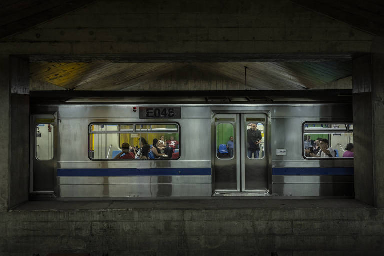 A imagem mostra um trem de metrô estacionado em uma estação subterrânea. O trem é prateado com uma faixa azul e possui janelas que revelam passageiros sentados e em pé. O ambiente é escuro, com um teto curvado e iluminação suave, criando um contraste com a estrutura de concreto da estação.