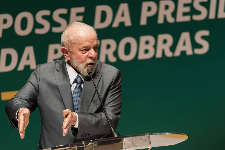 Imagem de um homem em um terno cinza, gesticulando enquanto fala em um evento. Ao fundo, há um painel verde com a frase 'POSSE DA PRESIDÊNCIA DA PETROBRAS'