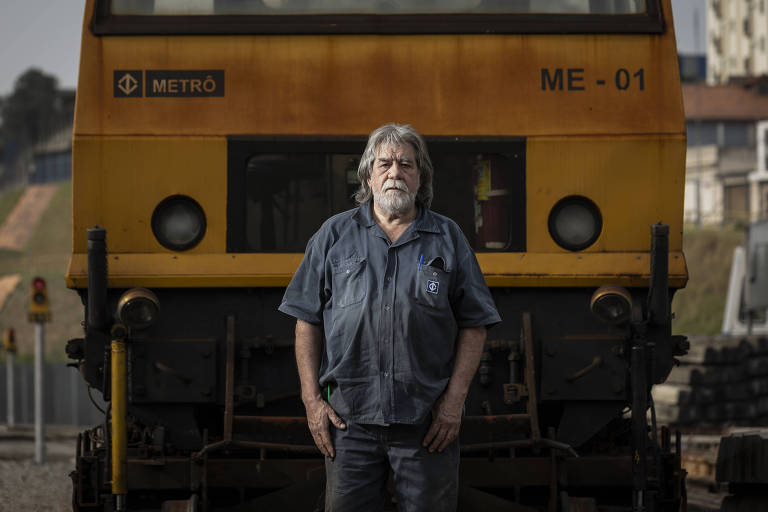 Um homem de cabelos longos e barba grisalha está em pé em frente a um trem amarelo e preto. Ele usa uma camisa azul e parece estar em um ambiente ferroviário, com trilhos e edifícios ao fundo. O trem tem a inscrição 'METRÔ' e o número 'ME - 01' visíveis.
