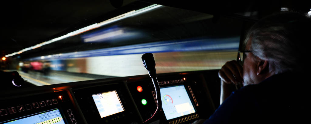 A imagem mostra a cabine de controle de um trem, com um operador visível à direita. O ambiente é escuro, com luzes de painel acesas, exibindo telas e botões de controle. O operador parece concentrado, observando a frente do trem enquanto ele se move.