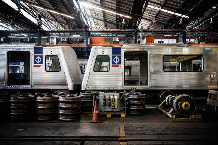 A imagem mostra o interior de uma oficina de manutenção de trens, com dois vagões de trem prateados estacionados. Os vagões têm logotipos visíveis na lateral. No chão, há várias rodas de trem empilhadas e um equipamento de manutenção em destaque. O ambiente é bem iluminado, com um teto alto e estrutura metálica.