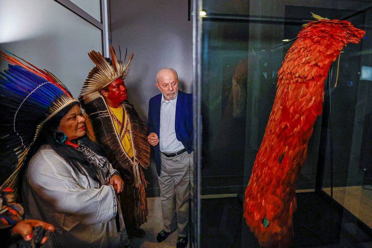 Presidente Lula observa o manto dos indígenas tupinambá, feito de penas vermelhas, ao lado de dois indígenas com trajes tradicionais e cocares de penas nas cabeças