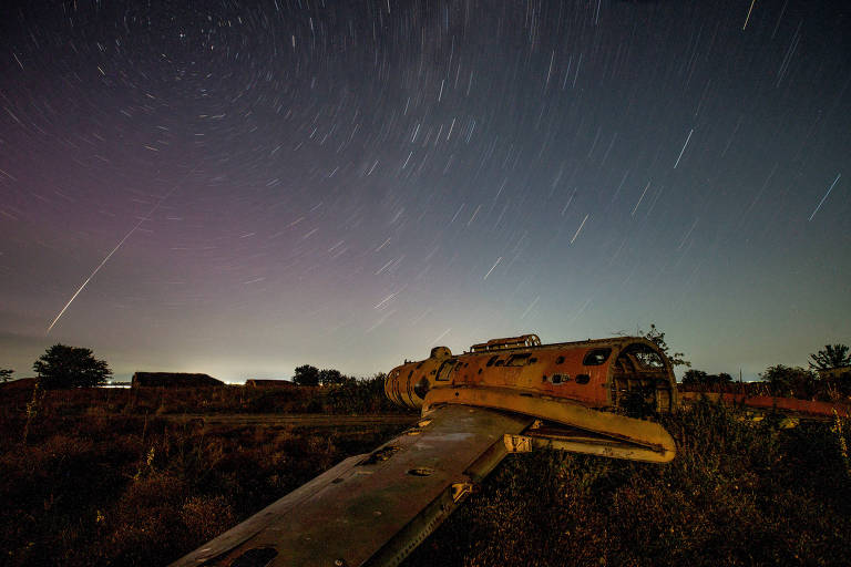 A imagem mostra um avião abandonado em um ambiente desértico sob um céu noturno estrelado. As estrelas estão visíveis em longas trilhas, indicando movimento, e há uma leve iluminação no horizonte. O avião está posicionado de lado, com a asa em primeiro plano e a fuselagem ao fundo, cercado por vegetação rasteira.
