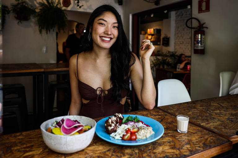 Uma mulher está sentada em uma mesa de madeira em um restaurante, sorrindo enquanto segura um garfo. À sua frente, há dois pratos: um prato azul com comida variada, incluindo arroz e vegetais, e uma tigela branca com frutas. Ao lado, há um pequeno copo com uma bebida. O ambiente é iluminado e decorado de forma moderna.