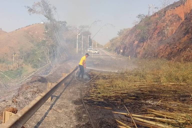 A imagem mostra um homem vestido com uma camiseta amarela, utilizando uma mangueira para combater um incêndio em uma área rural. Ao fundo, há uma estrada com alguns veículos e uma casa. O terreno é montanhoso, com vegetação queimada e fumaça visível no ar. O cenário é de um incêndio controlado, com a vegetação ao redor ainda em chamas