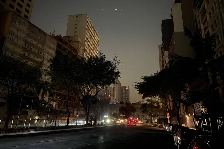 A imagem mostra uma rua urbana em uma noite escura, com prédios altos ao fundo. A iluminação é escassa, com algumas luzes de carros visíveis e árvores ao longo da calçada. O céu está quase sem estrelas e com uma camada de fumaça e poluição.