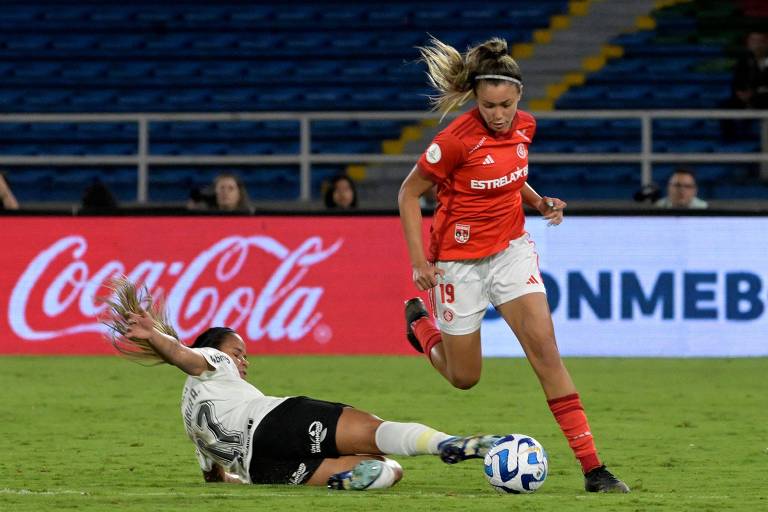 A imagem mostra uma jogadora de futebol em um campo, vestindo um uniforme vermelho com o número 19, correndo enquanto uma adversária, vestindo um uniforme branco e preto, está caída no chão. A jogadora no chão parece ter caído após uma disputa pela bola, que está próxima a ela. Ao fundo, há uma tela com o logotipo da Coca-Cola e arquibancadas vazias.
