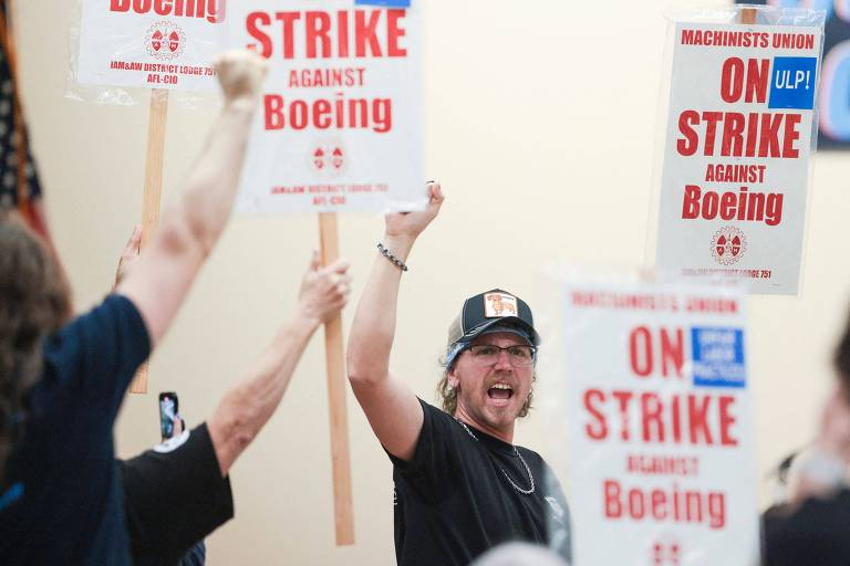 A imagem mostra um grupo de trabalhadores em um protesto, segurando cartazes que dizem 'Em greve' e 'Boeing'. Um dos manifestantes está levantando o punho em sinal de apoio. O ambiente parece ser interno, com uma parede clara ao fundo.