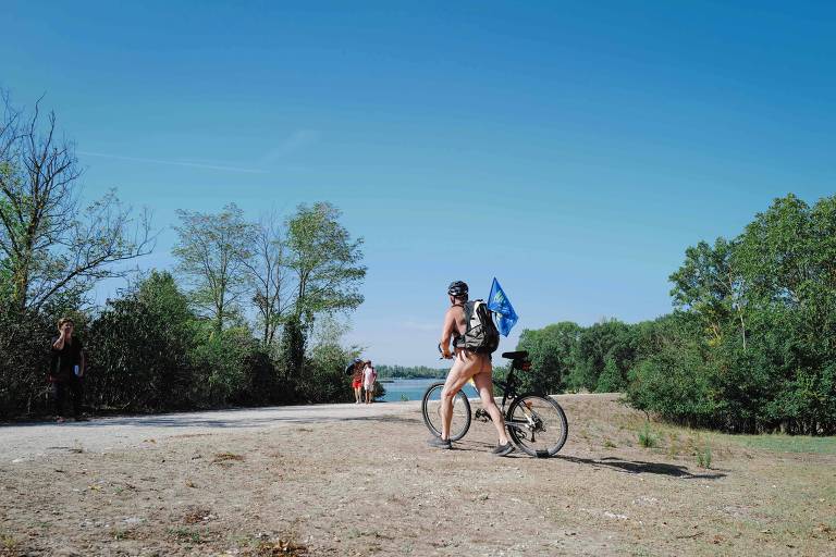 Um ciclista nu é visto em meio a uma paisagem de um parque. O dia é ensolarado, com céu azul e vegetação verde ao fundo.