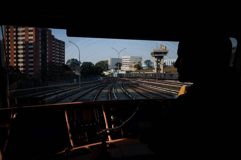 A imagem mostra a vista do interior de um trem, com o painel de controle visível em primeiro plano e o rosto de um homem a contraluz. No fundo, há uma paisagem urbana com prédios e uma torre de controle. A luz do sol ilumina a cena, criando um contraste entre a escuridão do interior do trem e a luminosidade externa.