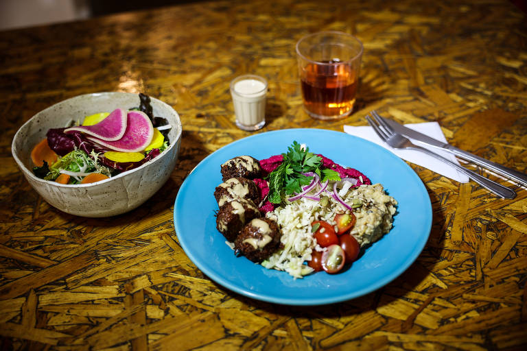 A imagem mostra um prato azul com carne grelhada, arroz, tomates-cereja e cebola roxa, decorado com folhas verdes. Ao lado, há uma tigela de salada com vegetais coloridos, incluindo fatias de beterraba e folhas verdes. Também estão presentes um copo de bebida laranja e um copo pequeno com um líquido claro, além de talheres em uma mesa de madeira.