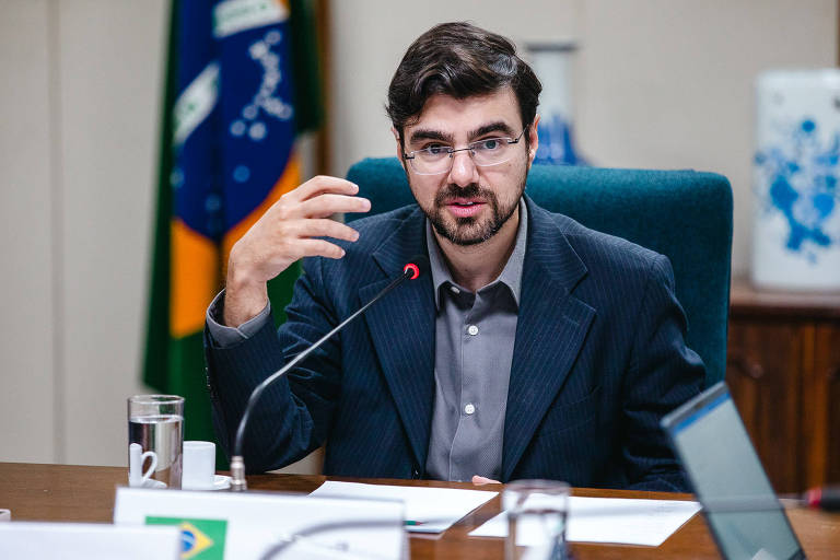 Secretário de Política Econômica do Ministério da Fazenda, Guilherme Mello, está sentado, veste um terno escuro com risca de giz, camisa cinza. Ele usa óculos e cavanhaque