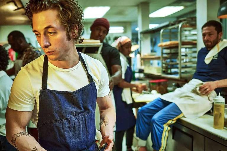 A foto mostra um homem jovem de pele e cabelos claros, com uma camiseta branca e um avental azul escuro em uma cozinha.
