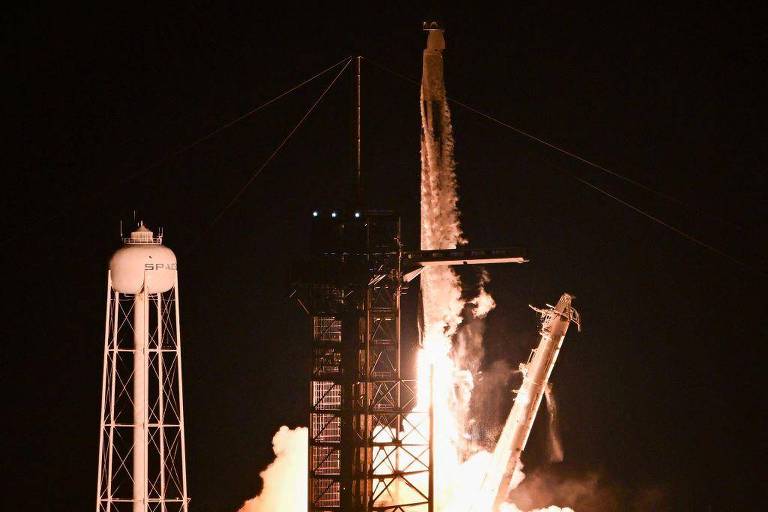 A imagem mostra um foguete sendo lançado à noite, com chamas e fumaça saindo da base. À esquerda, há uma torre de lançamento e um tanque de água com a inscrição 'SPACE'. O foguete está se elevando em direção ao céu, iluminando a cena ao seu redor.