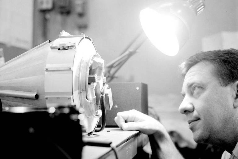 A imagem em preto e branco mostra um homem concentrado, olhando de perto para um equipamento técnico. O dispositivo tem uma forma cilíndrica e está iluminado por uma lâmpada de mesa. O ambiente parece ser um laboratório ou oficina, com outros equipamentos visíveis ao fundo.