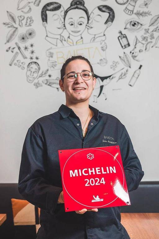 Uma pessoa sorridente está segurando um prêmio Michelin em um fundo com ilustrações. A pessoa usa uma camisa escura e parece estar em um ambiente de restaurante. O prêmio é redondo e vermelho, com a palavra 'MICHELIN' em destaque.