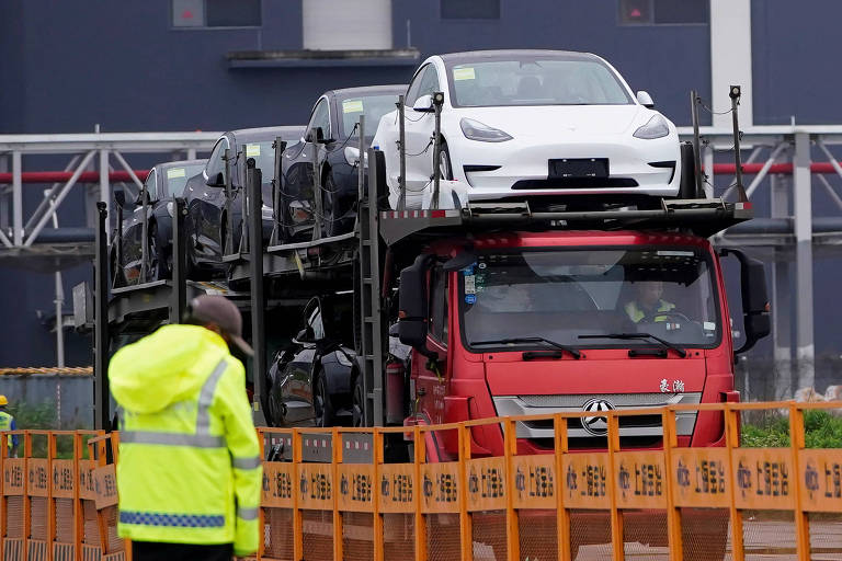 Um caminhão vermelho transporta vários carros, incluindo um modelo branco na parte superior. Ao fundo, há uma estrutura com o logotipo da Tesla. Um trabalhador vestido com um colete amarelo está em primeiro plano, observando o caminhão.