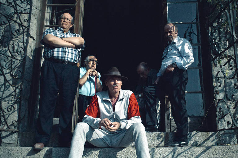 Cinco homens estão juntos olham para o horizonte. Ao centro, há um homem de camiseta branca e vermelha, com um chapéu preto. A sua esquerda, um homem em pé, de braços cruzados, usa uma camisa xadrez e outro está com uma bengala. A direita do homem de chapéu, há outro, sombreado, e mais um em pé.