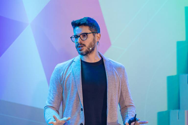 Fotografia colorida é retrato de Diogo Cortiz, homem branco de cabelos e barba castanhos. Veste camiseta preta e blazer cinza, usa óculos. Está falando em uma palestra