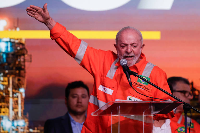 Foto mostra o presidente Luiz Inácio Lula da Silva, homem branco com um uniforme de trabalho laranja, em pé atrás de um púlpito de vidro, gesticulando com a mão levantada. Ao fundo, há uma imagem de uma instalação industrial. Um homem em um terno escuro está ao lado dele, observando