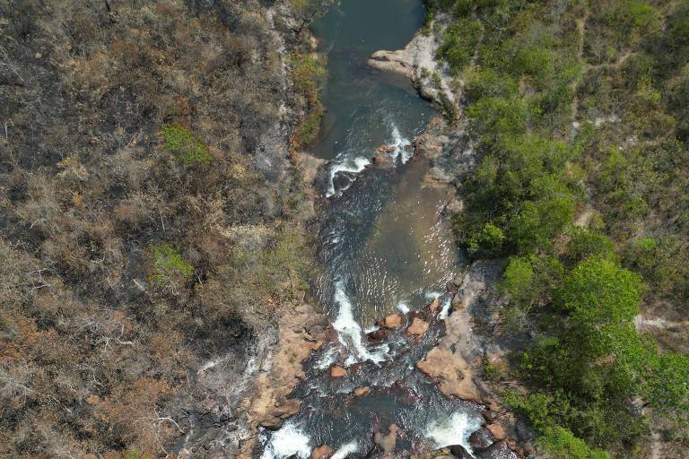 A imagem mostra uma vista aérea de um rio que flui entre rochas e vegetação. O rio é estreito e serpenteante, com áreas de água visíveis entre as pedras. A vegetação ao redor é predominantemente verde, com algumas áreas secas e expostas. Há uma mistura de rochas grandes e pequenas ao longo do leito do rio.