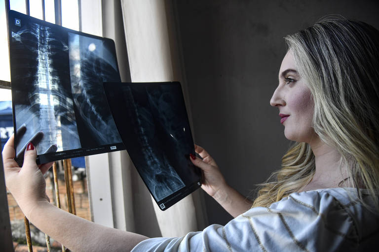 Uma mulher loura segura duas chapas de Raio-X diante de uma janela