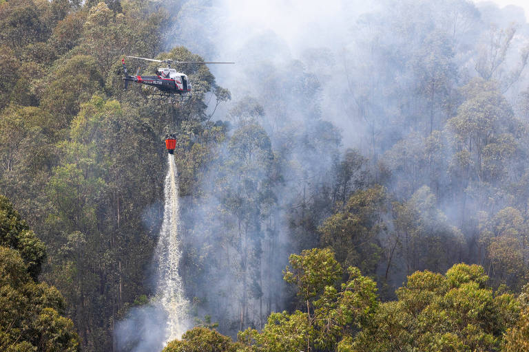 Um helicóptero sobrevoa uma área florestal densa, com fumaça visível ao redor. O helicóptero está despejando água em uma área afetada por incêndio, com um longo cabo conectado a um balde. A vegetação ao redor é verde, mas há sinais de fumaça que indicam a presença de fogo nas proximidades.
