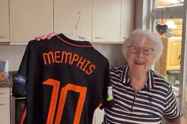       Uma mulher idosa, com cabelo branco e óculos, está sorrindo enquanto segura uma camisa de futebol preta com detalhes laranja. A camisa tem o nome 'MEMPHIS' e o número '10' nas costas. Ao fundo, pode-se ver uma cozinha com armários claros e utensílios.