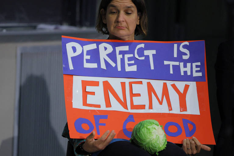 Uma mulher sentada segura um cartaz colorido que diz 'PERFECT IS THE ENEMY OF GOOD'. Ela parece pensativa e está segurando um repolho em seu colo. O cartaz é dividido em seções de cores laranja, roxa e branca, com letras grandes e destacadas.