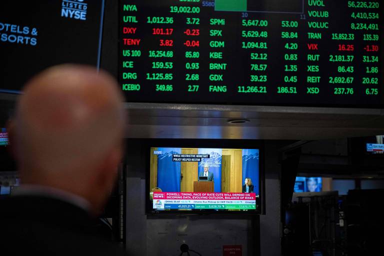 A imagem mostra um homem de costas, observando uma tela de televisão que transmite uma conferência. Na tela, um homem está em um pódio, enquanto uma mulher está sentada ao lado. Acima da tela, há uma lista de dados financeiros em verde e vermelho, indicando cotações de ações e índices. O ambiente parece ser a Bolsa de Valores, com informações financeiras visíveis.
