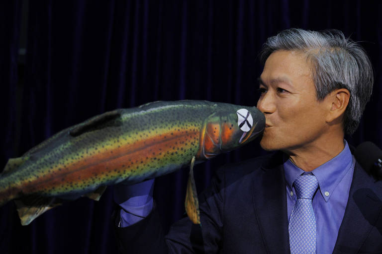 Um homem com cabelo grisalho e terno escuro está segurando um peixe colorido em sua mão, aproximando-o de seu rosto como se estivesse prestes a beijá-lo. O peixe tem uma coloração vibrante, com tons de verde, laranja e manchas pretas. O fundo é escuro, sugerindo um ambiente de apresentação ou conferência.