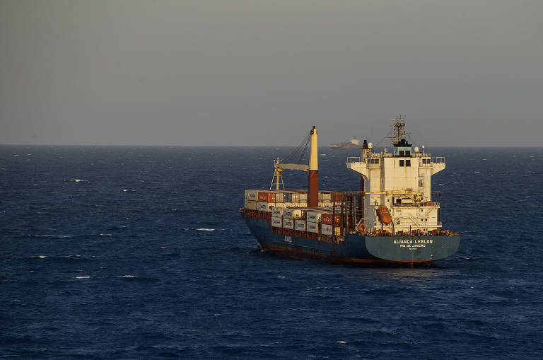 A imagem mostra um navio cargueiro navegando em águas abertas. O navio é de cor azul e possui várias estruturas， incluindo chaminés e guindastes. O céu está claro， e o mar parece calmo， com algumas ondas visíveis ao redor do navio.