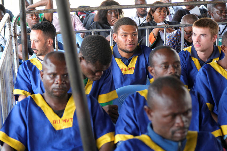 A imagem mostra um grupo de homens sentados em um ambiente fechado, todos vestindo uniformes azuis com detalhes amarelos. Alguns homens estão olhando para baixo, enquanto outros parecem estar conversando ou observando ao redor. O ambiente é bem iluminado e há uma grade visível ao fundo, sugerindo que estão em uma área restrita.