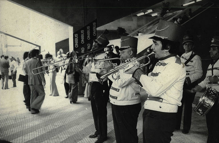 A imagem mostra uma banda de músicos em um evento público. Os músicos estão vestidos com uniformes brancos e chapéus vermelhos, tocando instrumentos como trombones e trompetes. Ao fundo, há um grupo de pessoas conversando e se movimentando. O ambiente parece ser um espaço amplo, possivelmente uma estação ou um centro de transporte, com escadas e placas visíveis.