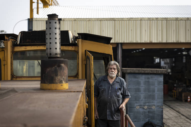 Um homem de cabelos grisalhos e barba, vestindo uma camisa escura, está em pé na entrada de uma locomotiva amarela. Ele parece estar em um ambiente industrial, com uma estrutura metálica ao fundo e um galpão coberto. A locomotiva tem um cano de escape visível e a cena é iluminada por luz natural.