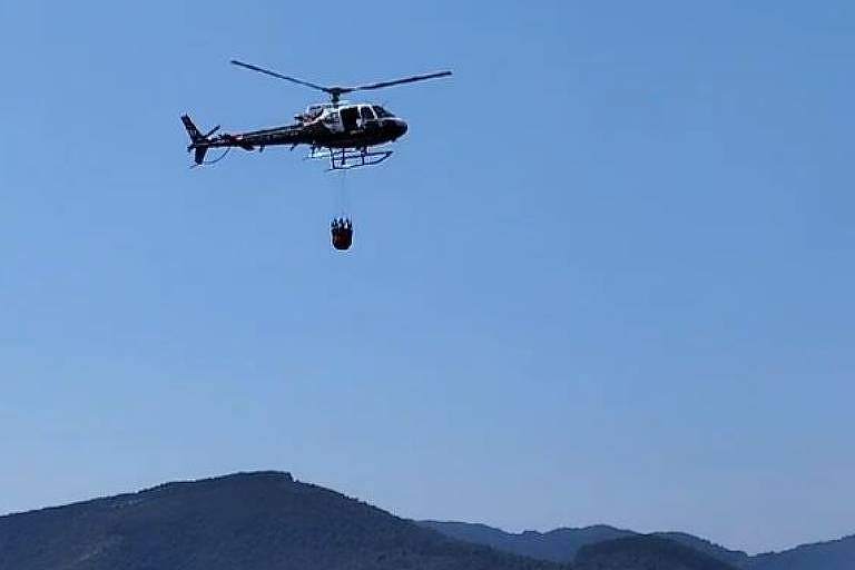 Helicóptero no ar, com um ceto pendurado, voa sobre um morro no céu azul