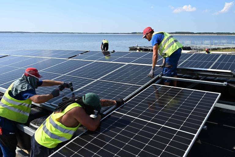 A imagem mostra trabalhadores instalando painéis solares em uma plataforma flutuante sobre a água. Quatro homens estão envolvidos na instalação, usando coletes refletivos e chapéus. Um dos trabalhadores está usando uma furadeira, enquanto outro ajusta um painel. Ao fundo, vê-se um lago e uma área verde.
