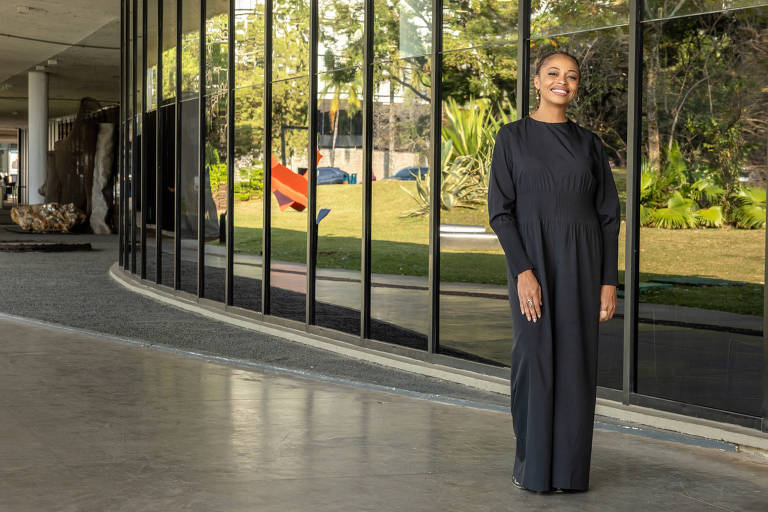 Uma mulher está posando em um ambiente moderno com grandes janelas de vidro. Ela usa um vestido longo e escuro, e sorri enquanto se apoia levemente em uma das mãos. Ao fundo, há um jardim com plantas e esculturas, refletindo a luz do sol nas janelas.