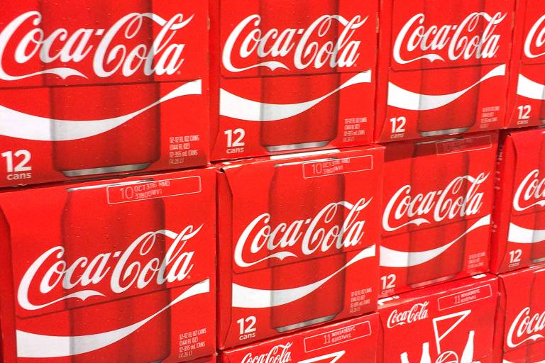 A imagem mostra várias caixas de refrigerante Coca-Cola empilhadas. Cada caixa é vermelha com o logotipo da Coca-Cola em branco. As caixas contêm 12 latas cada e estão organizadas em uma prateleira. Algumas informações de data e código de barras são visíveis nas caixas.
