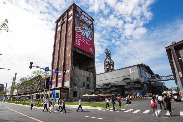 A imagem mostra um edifício moderno e alto, com uma grande faixa publicitária na parte superior. Na frente do edifício, várias pessoas estão atravessando a rua. O céu está parcialmente nublado, com algumas nuvens visíveis. O ambiente urbano é bem iluminado e há um tráfego de veículos visível na rua.