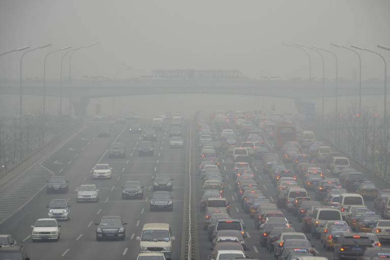 A imagem mostra uma estrada com intenso tráfego de veículos, visível em meio a uma densa neblina. Os carros estão parados em várias faixas, e a visibilidade é bastante reduzida devido à poluição do ar. Ao fundo, há uma ponte e postes de iluminação ao longo da estrada