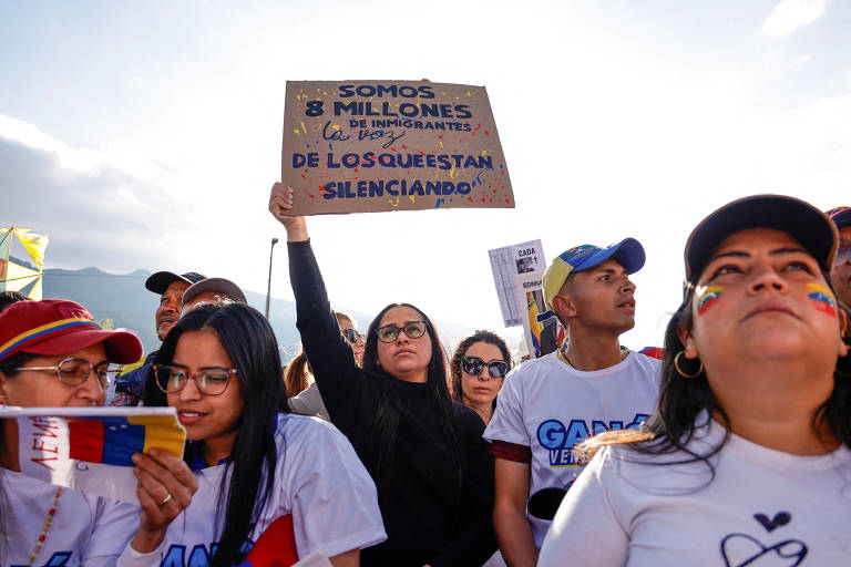 A imagem mostra um grupo de pessoas participando de um protesto. Uma mulher em destaque segura um cartaz que diz: 'SOMOS 8 MILLONES DE INMIGRANTES LA VOZ DE LOS QUE ESTÁN SILENCIANDO'. As pessoas ao redor estão vestindo camisetas brancas e algumas têm bandeiras venezuelanas pintadas no rosto. O cenário é ao ar livre, com montanhas ao fundo e um céu parcialmente nublado.
