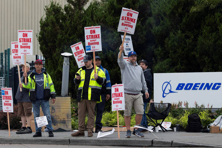 Um grupo de trabalhadores em greve está em frente à fábrica da Boeing, segurando cartazes que dizem 'ON STRIKE AGAINST Boeing'. Os manifestantes estão vestidos com coletes de segurança e alguns usam bonés. Um dos cartazes adicionais diz 'WE ARE NOT LAZY OR GREEDY'. Ao fundo, há um letreiro da Boeing.
