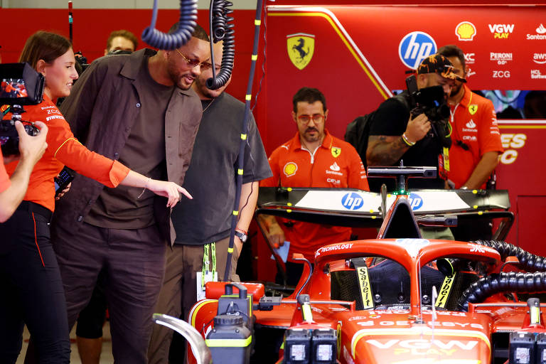 À esquerda mulher de camisa vermelha aponta para carro de F1 parado no boxe da Ferrari