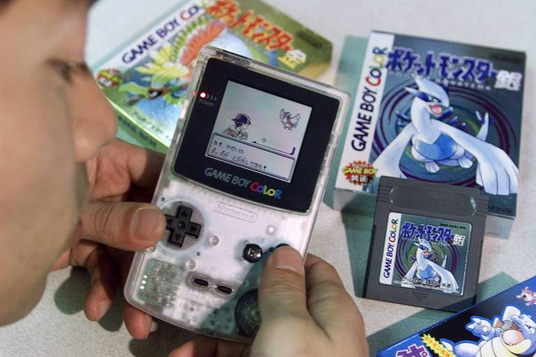 A imagem mostra uma pessoa segurando um Game Boy Color transparente enquanto joga. Na tela do console, é possível ver um jogo de Pokémon, com um personagem e um Pokémon em batalha. Ao fundo, há caixas de jogos de Pokémon e um cartucho visível ao lado do console.
