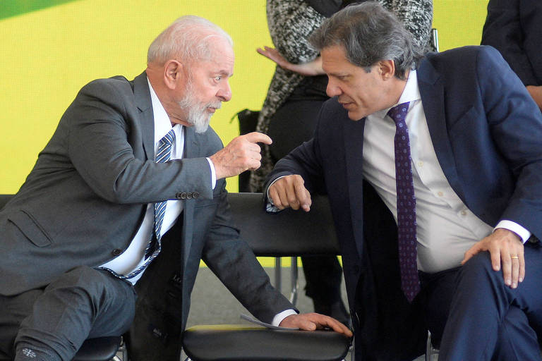 Dois homens sentados em cadeiras, conversando de forma animada. Um deles, com cabelo grisalho e barba, usa um terno escuro e gesticula com a mão. O outro, com cabelo escuro e terno claro, escuta atentamente. Ao fundo, uma parede verde e pessoas em pé.