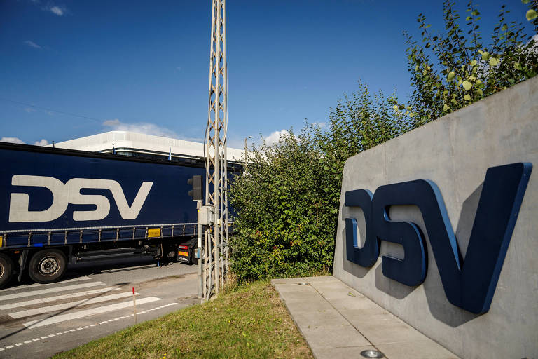 A imagem mostra um caminhão azul da DSV estacionado próximo a um letreiro da empresa, que é feito de concreto e apresenta as letras 'DSV' em destaque. Ao fundo, há uma estrutura coberta e vegetação ao redor, sob um céu claro.
