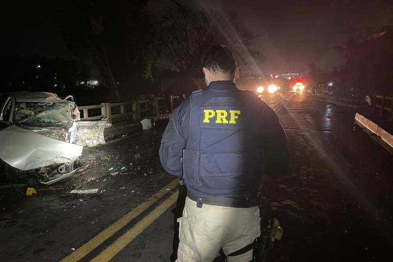 A imagem mostra um agente da Polícia Rodoviária Federal (PRF) de costas, usando um colete azul com a sigla 'PRF' em amarelo. Ao fundo, há um carro acidentado na pista, com destroços visíveis. A cena é iluminada por luzes de viaturas, indicando que é à noite. A estrada apresenta marcas de pneus e está parcialmente bloqueada.