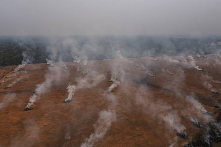 Imagem aérea de uma área de terra queimada, com fumaça subindo em várias partes. O solo é de cor marrom claro, e ao fundo, há uma densa vegetação verde, indicando a presença de floresta. A fumaça se espalha pelo ar, criando uma atmosfera de poluição.